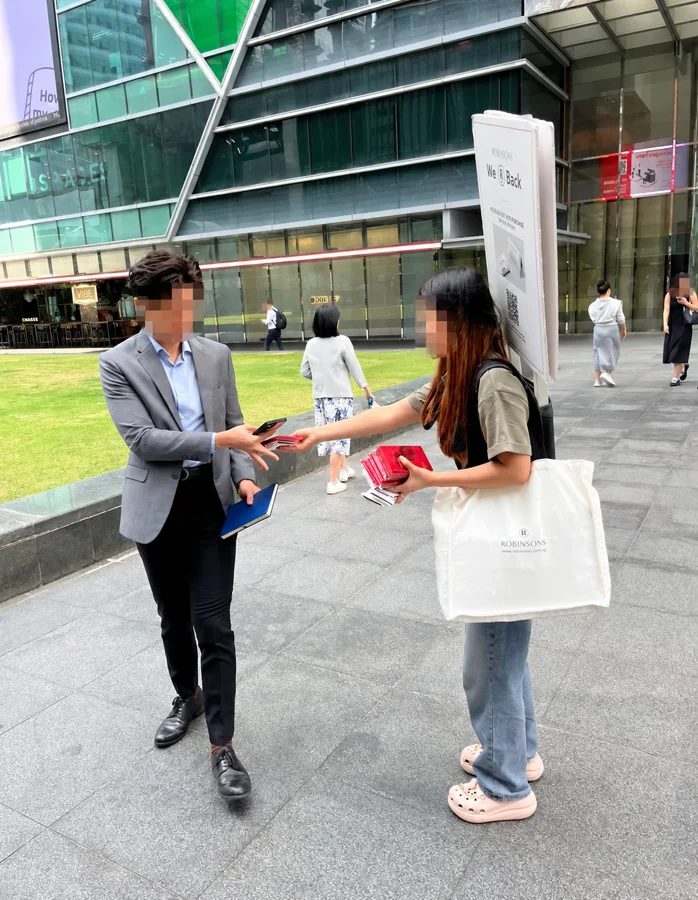 Ang bao packets and promotional tissue packs distributed during a Robinsons street campaign, supported by a backpack advertising board.
